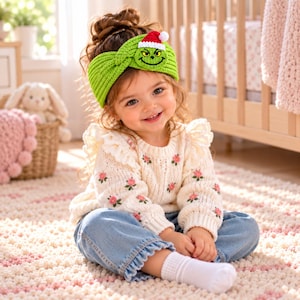 Könnte beinhalten: Ein leuchtend grünes Strickstirnband mit einem gehäkelten Grinch-Gesicht und einer kleinen rot-weißen Weihnachtsmütze. Das Stirnband hat ein gedrehtes Knotendetail auf der Vorderseite und wird zu einem cremefarbenen Langarmpullover mit rosa Blumenstickerei getragen.