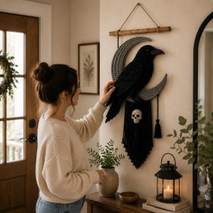 May include: A wall hanging featuring a black macrame raven perched on a grey crescent moon. The piece is suspended from a wooden dowel with a rope. A small white skull charm hangs from the bottom of the raven, followed by black macrame fringe.