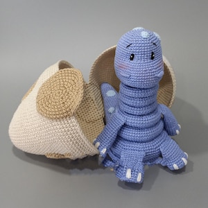 May include: A crocheted blue dinosaur toy with white accents, emerging from a tan and cream-colored eggshell. The dinosaur has a long neck, small arms and legs, and a friendly expression. The eggshell is open, revealing the dinosaur.