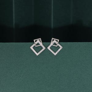 Puede incluir: Un par de pendientes de botón plateados. Cada pendiente presenta dos formas cuadradas superpuestas, la exterior incrustada con pequeñas piedras brillantes. Los pendientes se muestran sobre una superficie verde oscuro.