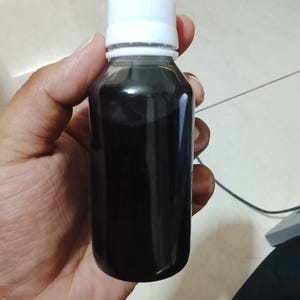 May include: A small, clear plastic bottle filled with a dark, opaque liquid. The container features a smooth, cylindrical body and is sealed with a simple, ribbed white screw-on cap. The liquid inside appears completely black and non-transparent.
