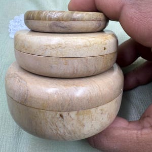 Può includere: Tre scatole rotonde in pietra beige impilate con coperchi. Le scatole sono di dimensioni diverse, la più piccola in cima. La pietra ha un aspetto naturale marmorizzato con toni crema e marrone chiaro. Le scatole sono tenute da una mano.