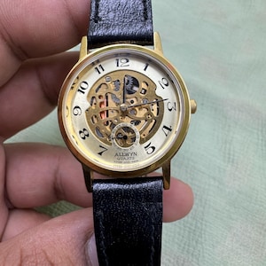 Op de afbeelding: Een goudkleurig Allwyn Quartz horloge met een zwarte leren band. De wijzerplaat van het horloge is crèmekleurig met zwarte cijfers en een gedeeltelijk zichtbaar skeleton design. Het horloge wordt door een hand vastgehouden.