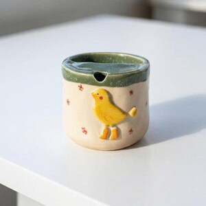 May include: A ceramic ashtray with a yellow bird design. The ashtray is cylindrical with a green rim and a small opening. The bird is wearing orange boots and is on a cream-colored background with red floral accents.