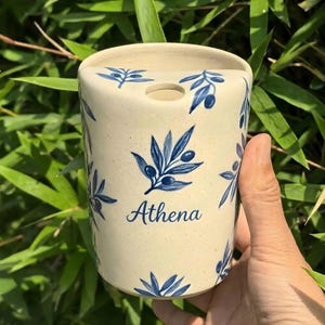May include: A cylindrical ceramic container with a cream-colored base, adorned with blue olive branch designs. The word "Athena" is written in blue script. The container has a hole at the top.