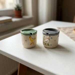 May include: Two ceramic cups with a farm theme. One cup has a yellow bird, the other a white goose. Both cups have a green or gray rim and small floral accents. The cups are on a white surface.