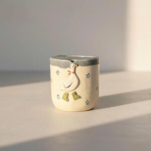 May include: A cream-colored ceramic mug with a whimsical goose design. The goose has a red bow and green feet. Blue paw prints decorate the mug's surface. The rim is gray.