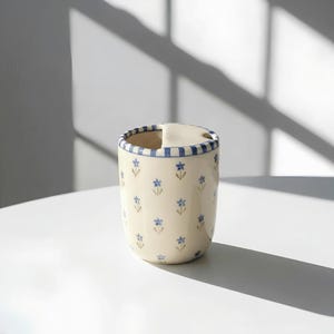 Könnte beinhalten: Ein cremefarbener Keramikbehälter mit blau gestreiftem Rand und kleinen blauen Blumenmustern. Der Behälter hat eine eingebaute Löffelablage und dient wahrscheinlich zur Aufbewahrung in der Küche oder als Utensilienhalter. Höhe ca. 12 cm.