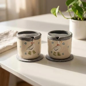 May include: Two ceramic containers with a duck design. The containers are off-white with a gray rim and a duck illustration with a red bow. Blue floral accents are scattered around the duck. The containers sit on gray felt coasters.
