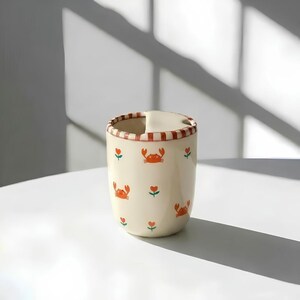 May include: A cream-colored ceramic mug with a crab and flower pattern. The mug has a red and white striped rim and a small handle. The crab illustrations are orange, and the flowers are red with green stems. The mug is on a white surface.