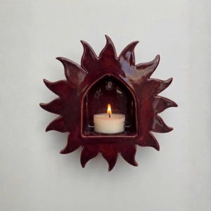 May include: A burgundy ceramic sun-shaped wall sconce with a small arched alcove holding a lit white tea light candle. The sconce has pointed rays around the perimeter and is mounted on a white wall.