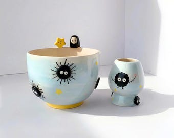 Pode incluir: Uma tigela de cerâmica azul-clara e uma chávena mais pequena a condizer, ambas com personagens pretas de fuligem, estrelas amarelas e uma pequena figura de rosto branco no rebordo da tigela. A tigela tem uma base amarela e forma redonda.