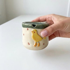May include: A ceramic ashtray with a yellow bird design. The ashtray is cylindrical with a green rim and a small opening. The bird is in relief, with orange boots and a red beak. Small red flower accents are scattered around the sides.