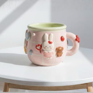 Peut inclure: Une tasse en céramique rose avec un intérieur vert clair et une anse. La tasse est décorée de personnages de dessins animés en 3D, dont un lapin, un ours en peluche et un ballon en forme de cœur. La tasse a un motif quadrillé.