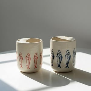 Puede incluir: Dos tazas de cerámica blanquecina con forma cilíndrica. Una taza presenta ilustraciones de peces rojos, mientras que la otra tiene diseños de peces azules. Las tazas están sobre una superficie blanca, con sombras.