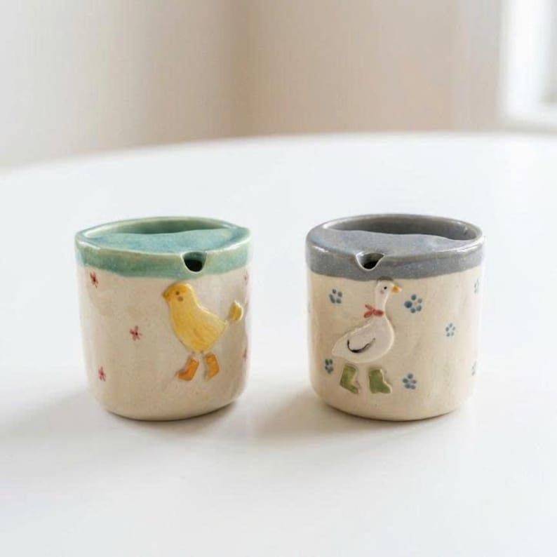 May include: Two ceramic ashtrays with a whimsical design. One features a yellow bird, the other a white goose with a red bow. Both have a light blue or gray rim and are decorated with small flowers and blue footprints.