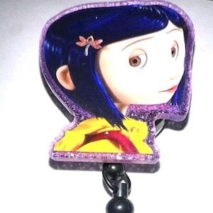 May include: A Coraline-themed badge reel featuring a character image with blue hair and a yellow raincoat. The reel includes a purple glittery border, a black bead, a purple rhinestone bead, and a yellow bead.