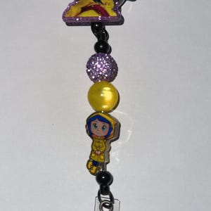 May include: A retractable badge reel featuring a cartoon character with blue hair and a yellow coat. The reel includes purple and yellow beads, and a clear plastic holder with a pink glitter accent.