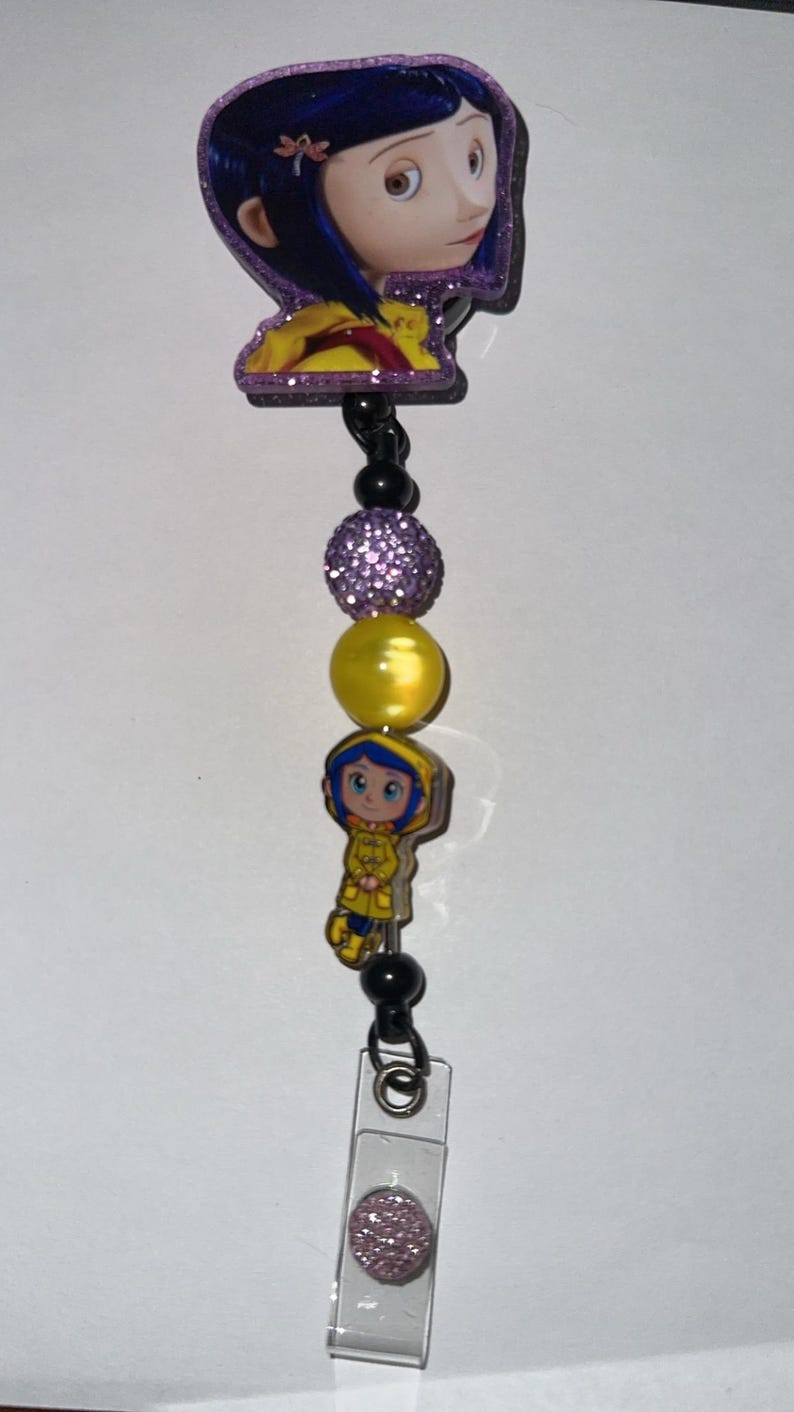 May include: A retractable badge reel featuring a cartoon character with blue hair and a yellow coat. The reel includes purple and yellow beads, and a clear plastic holder with a pink glitter accent.