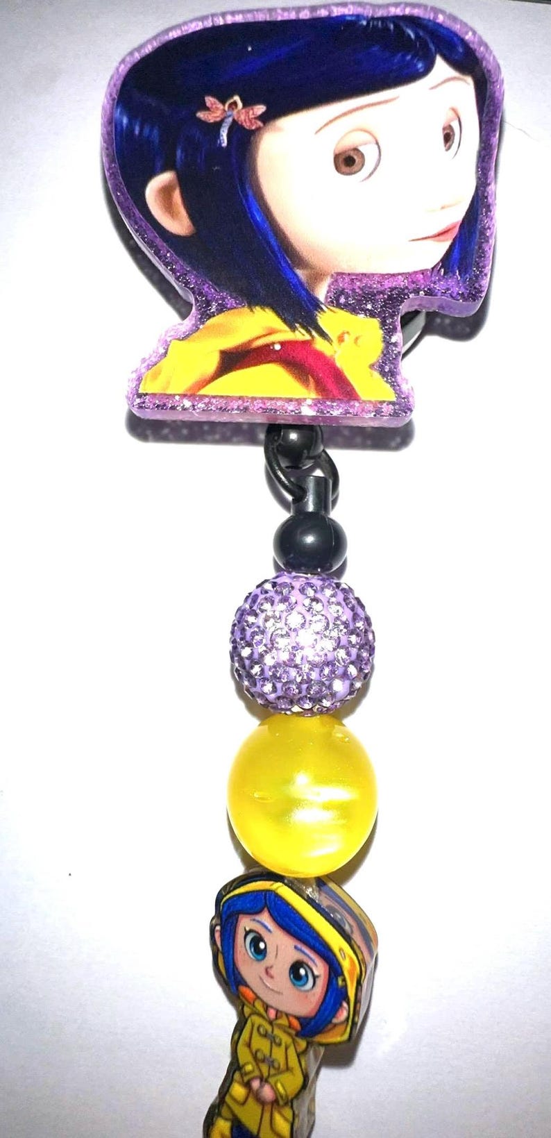 May include: A Coraline-themed badge reel featuring a character image with blue hair and a yellow raincoat. The reel includes a purple glittery border, a black bead, a purple rhinestone bead, and a yellow bead.