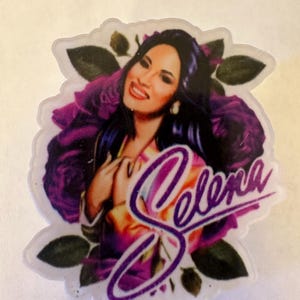May include: A badge reel featuring a portrait of Selena, surrounded by purple roses and green leaves. The name "Selena" is written in a cursive font over the image. The reel has a purple retractable cord and a clip.