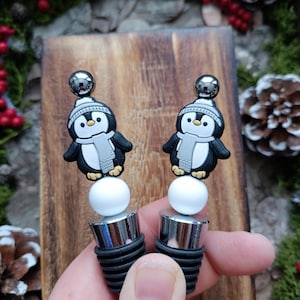 Könnte beinhalten: Zwei Flaschenverschlüsse mit Pinguin-Motiv. Jeder Verschluss zeigt einen schwarz-weißen Pinguin mit grauem Schal und Hut, der auf einer weißen Perle und einem schwarzen Gummistopfen sitzt. Die Verschlüsse haben einen silbernen Metallaufsatz.