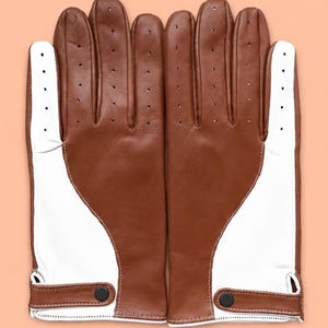 May include: A pair of driving gloves featuring a two-tone design with brown leather on the back and white leather on the palm side. The gloves include perforated detailing along the fingers, white contrast stitching, and a brown leather wrist strap.
