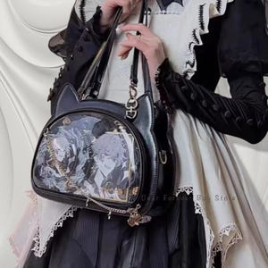 May include: Black handbag with cat ear accents and a clear front panel displaying anime-style character art. The bag features a chain strap, a top handle, and decorative charms. The bag is held against a white and black outfit.