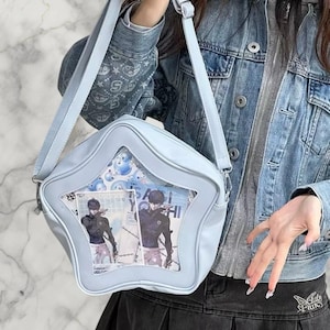 Puede incluir: Bolso de hombro azul claro en forma de estrella con una ventana transparente que muestra ilustraciones de estilo anime. El bolso tiene una correa larga y un cierre de solapa. Hecho de material liso, posiblemente polipiel.