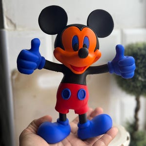 Pu&ograve; includere: Una statuetta di Topolino con orecchie, testa e braccia nere, viso arancione e occhi blu. La figura indossa pantaloncini rossi con bottoni blu, guanti blu e scarpe blu. La figura fa il pollice in su con entrambe le mani.