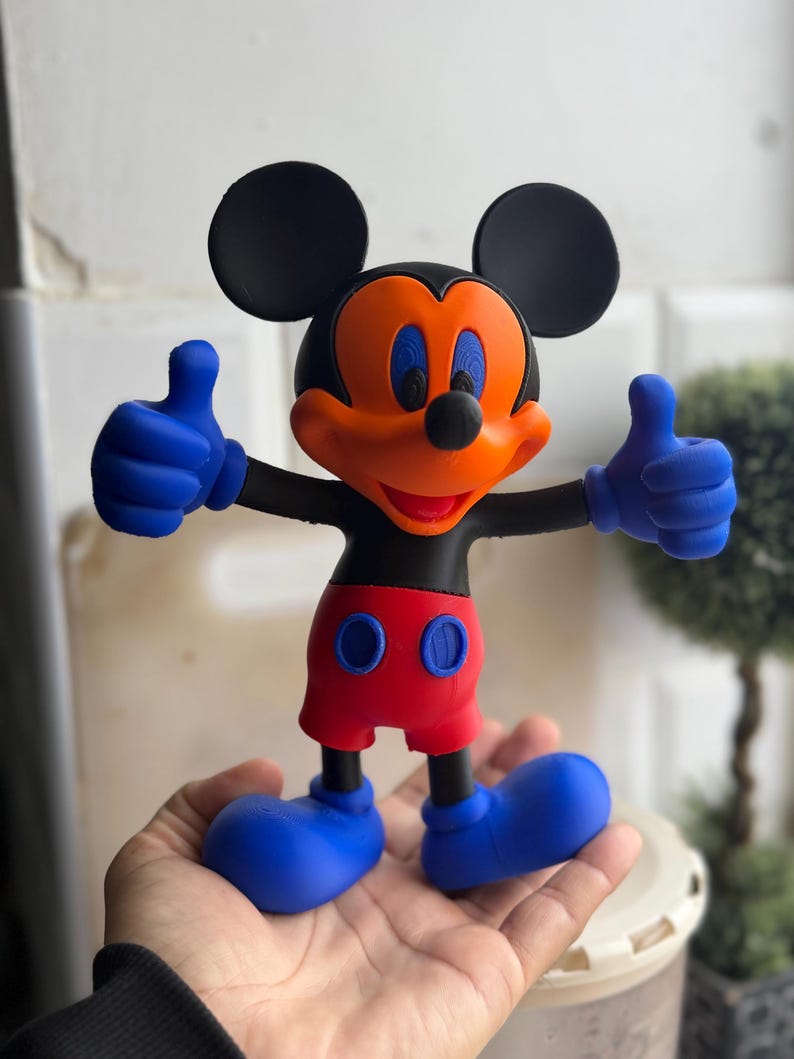 Pu&ograve; includere: Una statuetta di Topolino con orecchie, testa e braccia nere, viso arancione e occhi blu. La figura indossa pantaloncini rossi con bottoni blu, guanti blu e scarpe blu. La figura fa il pollice in su con entrambe le mani.