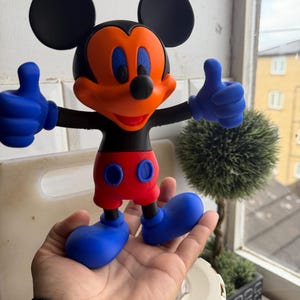 Pu&ograve; includere: Una colorata statuetta di Topolino con orecchie nere, viso arancione e occhi blu. La figura ha guanti blu che fanno il pollice in su, pantaloncini rossi e scarpe blu. La figura &egrave; tenuta in una mano.