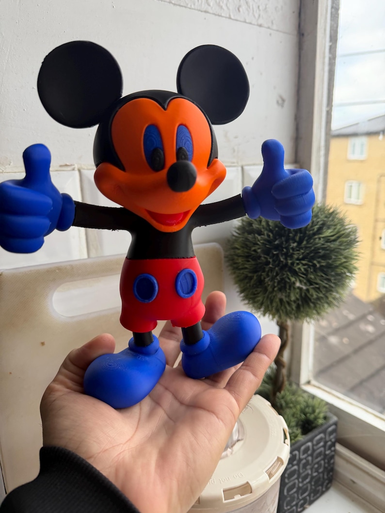 Pu&ograve; includere: Una colorata statuetta di Topolino con orecchie nere, viso arancione e occhi blu. La figura ha guanti blu che fanno il pollice in su, pantaloncini rossi e scarpe blu. La figura &egrave; tenuta in una mano.
