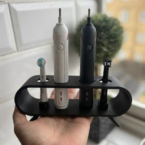 Puede incluir: Un soporte negro para cepillos de dientes con cuatro ranuras, que contiene dos cepillos de dientes eléctricos y dos cabezales. Un cepillo de dientes es blanco, el otro es negro. El soporte se sostiene en una mano.