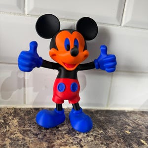 Pu&ograve; includere: Una colorata statuetta di Topolino con orecchie nere, viso arancione e bocca rossa. La figura ha guanti, scarpe blu e pantaloncini rossi. La figura &egrave; in posa con entrambi i pollici in su.