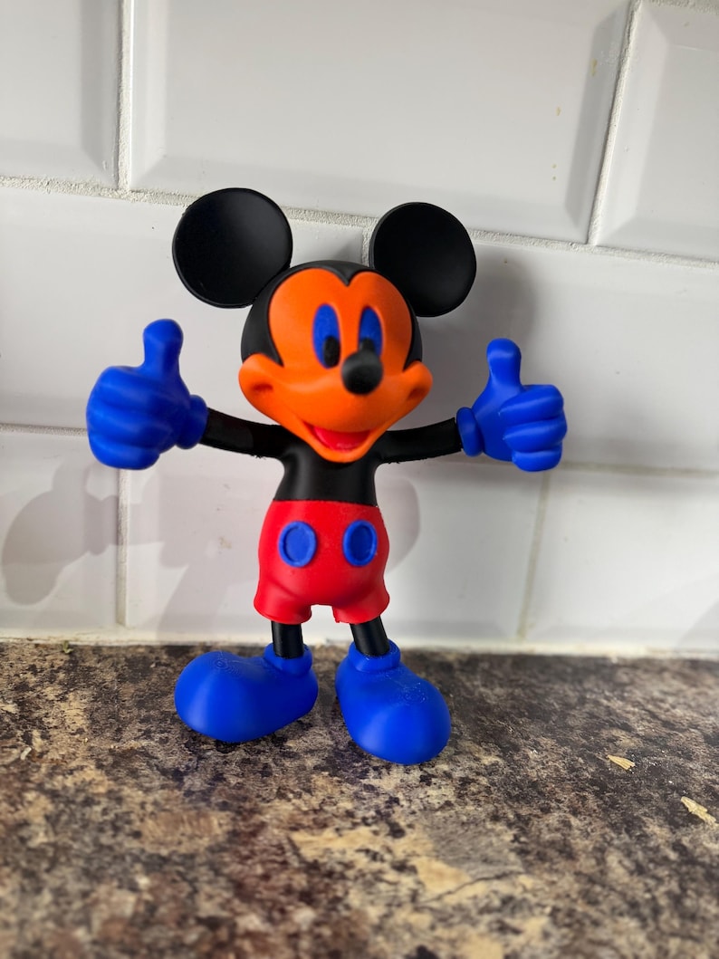 Pu&ograve; includere: Una colorata statuetta di Topolino con orecchie nere, viso arancione e bocca rossa. La figura ha guanti, scarpe blu e pantaloncini rossi. La figura &egrave; in posa con entrambi i pollici in su.