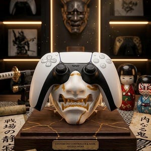 May include: A white and black gaming controller sits above a white and gold Oni mask, presented on a wooden base. The base has gold detailing and a plaque reading "CUSTOM ONI CONTROLLER & MASK ARTISAN WORK". The background includes a shelf with other controllers and Japanese-themed items.