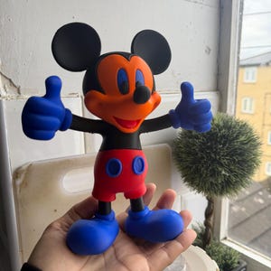 Pu&ograve; includere: Una statuetta di Topolino con orecchie nere, viso arancione e pantaloncini rossi. La figura ha guanti e scarpe blu e fa il gesto del pollice in su con entrambe le mani. Un personaggio popolare della Disney.