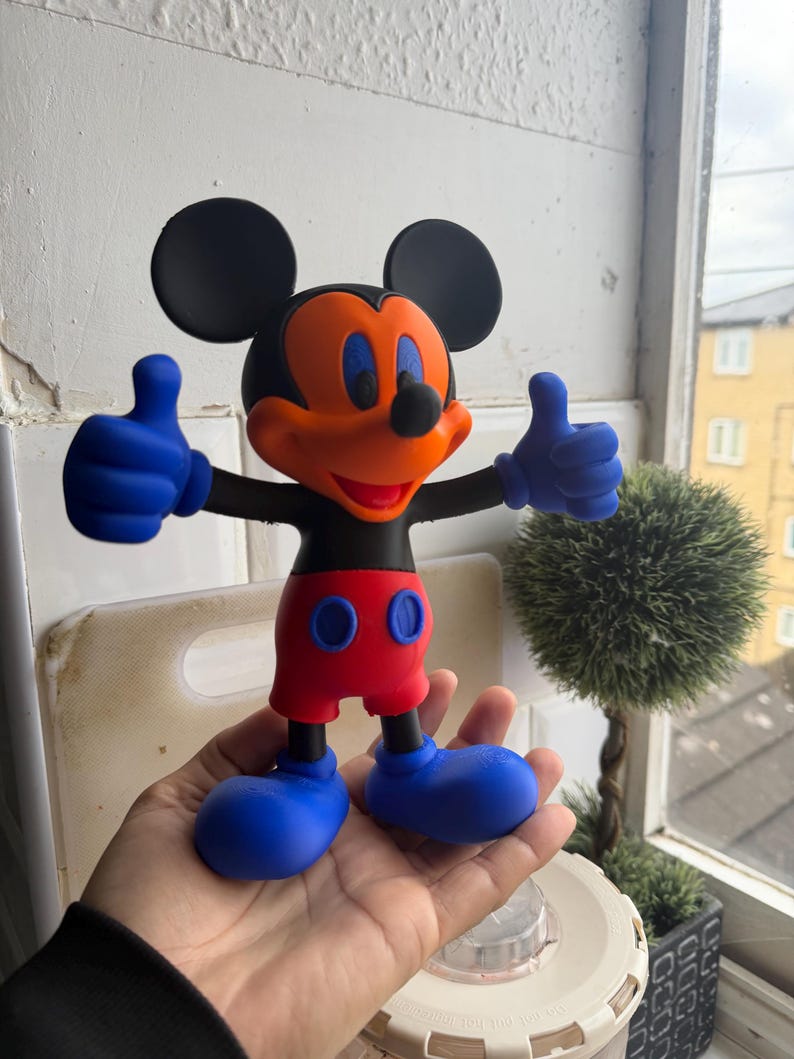 Pu&ograve; includere: Una statuetta di Topolino con orecchie nere, viso arancione e pantaloncini rossi. La figura ha guanti e scarpe blu e fa il gesto del pollice in su con entrambe le mani. Un personaggio popolare della Disney.