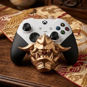 May include: A white Xbox controller with black grips and buttons, paired with a gold-colored Oni mask. The controller features colored buttons and a directional pad. The mask is intricately detailed and adds a unique aesthetic.