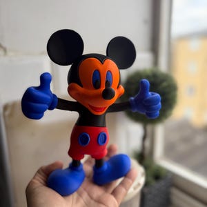Pu&ograve; includere: Una colorata statuetta di Topolino con orecchie nere, testa nera e viso arancione. La statuetta ha mani e piedi blu, pantaloncini rossi e fa il gesto del pollice in su. La statuetta &egrave; alta circa 15 cm.