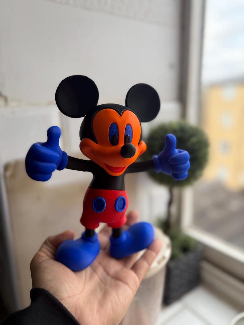 Pu&ograve; includere: Una colorata statuetta di Topolino con orecchie nere, testa nera e viso arancione. La statuetta ha mani e piedi blu, pantaloncini rossi e fa il gesto del pollice in su. La statuetta &egrave; alta circa 15 cm.