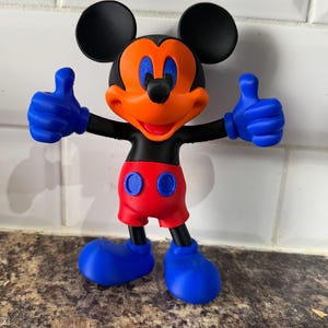 Pu&ograve; includere: Una colorata statuetta di Topolino con orecchie nere, corpo nero e viso arancione. La figura ha guanti e scarpe blu, pantaloncini rossi e fa il gesto del pollice in su. La figura &egrave; su una superficie marrone.