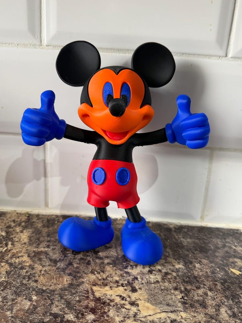 Pu&ograve; includere: Una colorata statuetta di Topolino con orecchie nere, corpo nero e viso arancione. La figura ha guanti e scarpe blu, pantaloncini rossi e fa il gesto del pollice in su. La figura &egrave; su una superficie marrone.