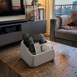 Puede incluir: Un organizador de control remoto de tela gris sobre una mesa de centro de madera rústica. El organizador contiene varios controles remotos negros, un controlador de juegos y un pequeño altavoz inteligente. Un televisor y una estantería están en el fondo.
