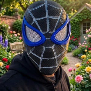 Puede incluir: Una m&aacute;scara negra de Spider-Man con un dise&ntilde;o de telara&ntilde;a y contornos de ojos azules. La m&aacute;scara tiene una superficie texturizada y la lleva una persona con una sudadera con capucha negra. El fondo muestra un jard&iacute;n con flores y un peque&ntilde;o edificio.
