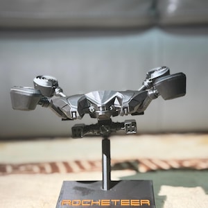 May include: A dark grey model of a futuristic aircraft, mounted on a black stand. The model features intricate geometric designs and is labelled "ROCKETEER ARC Raiders" in orange lettering on the base. The aircraft has wing-like structures and multiple engine components.