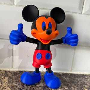 Pu&ograve; includere: Una statuetta di Topolino con orecchie nere, corpo nero e viso arancione brillante. La figura indossa pantaloncini rossi con bottoni blu, guanti blu e scarpe blu. Mickey alza i pollici con entrambe le mani.