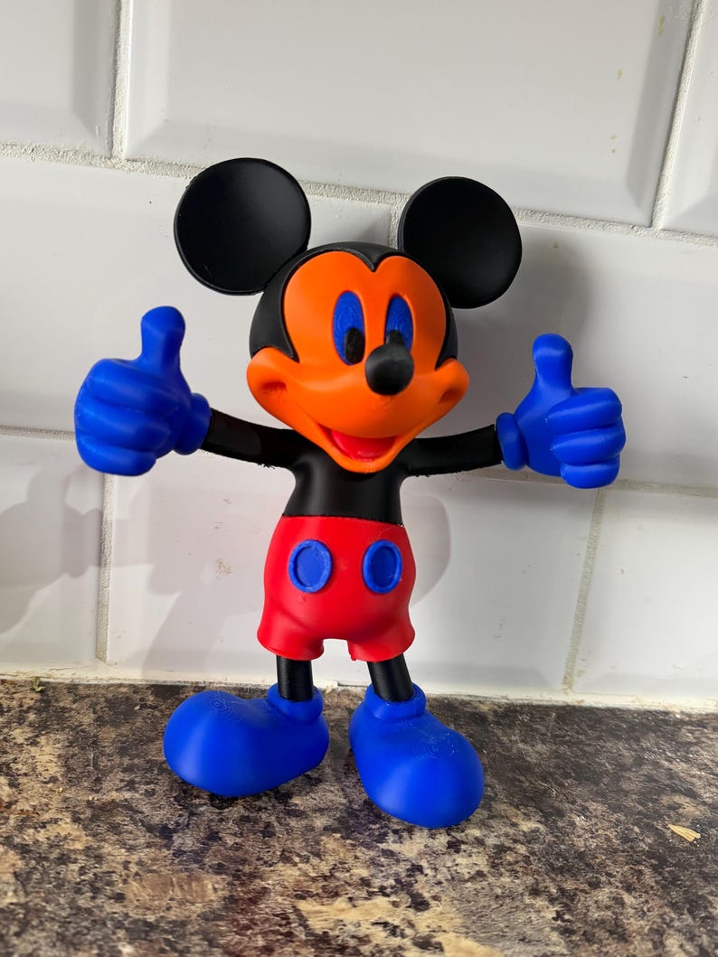 Pu&ograve; includere: Una statuetta di Topolino con orecchie nere, corpo nero e viso arancione brillante. La figura indossa pantaloncini rossi con bottoni blu, guanti blu e scarpe blu. Mickey alza i pollici con entrambe le mani.