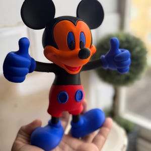 Pu&ograve; includere: Una colorata statuina di Topolino, con orecchie nere, corpo nero, viso arancione, occhi blu, pantaloncini rossi e guanti e scarpe blu. La figura fa il gesto del pollice in su con entrambe le mani. La statuina &egrave; tenuta in una mano.